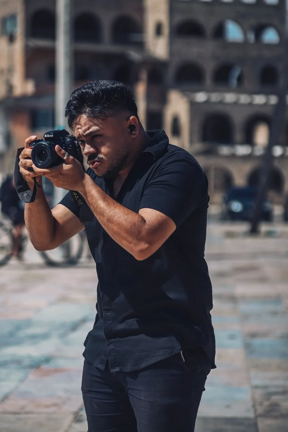 Johnny, de camisa preta, segurando uma câmera Nikon junto ao rosto em uma praça.
